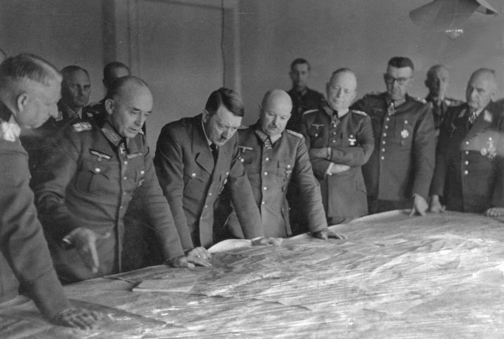 Hitler podczas narady z oficerami na froncie Wschodnim. Generał Zeitzler (z wąsem) stoi obok Hitlera (domena publiczna).