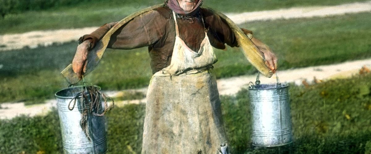 Wiejska kobieta niosąca wodę z użyciem koromysła.